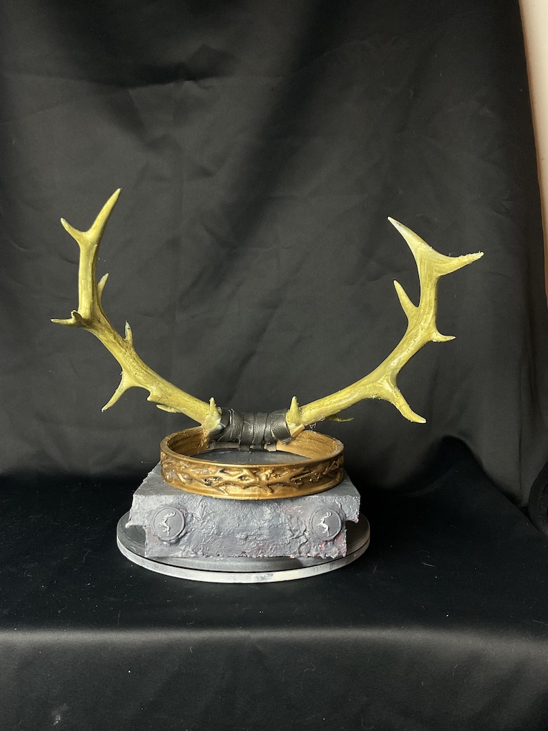 May include: A golden-colored antler crown with a decorative band and a gray base. The crown features a detailed design and is likely a costume piece or decorative item. The antlers are attached to the band with a dark material.