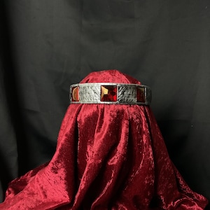 Puede incluir: Una diadema metálica con un acabado plateado texturizado y tres piedras translúcidas de color rojo oscuro de corte cuadrado engastadas a lo largo de la banda frontal. La piedra central está flanqueada por dos piedras rojas cuadradas más pequeñas.