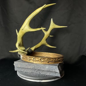 May include: A decorative antler sculpture with a gold-colored crown and a gray base. The antlers are a light green color. The crown is adorned with a detailed pattern. The base is made of stacked gray blocks.