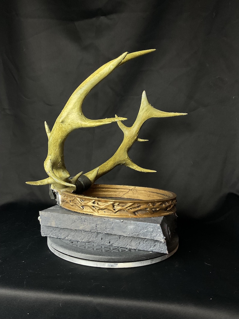 May include: A decorative antler sculpture with a gold-colored crown and a gray base. The antlers are a light green color. The crown is adorned with a detailed pattern. The base is made of stacked gray blocks.