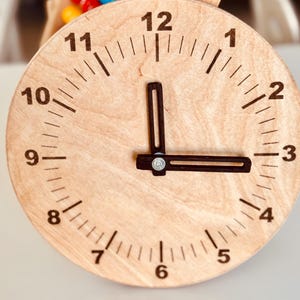 May include: A wooden clock face with black hands and black numbers. The clock face is light brown with a wood grain pattern. The numbers 1 through 12 are displayed around the clock face.
