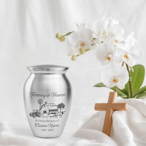 May include: A silver cremation urn with a farm scene engraving, including a tractor, barn, and animals. The text reads "Farming in Heaven" and "In Loving Memory of Custom Name 1988-2025". A wooden cross and white flowers are in the background.