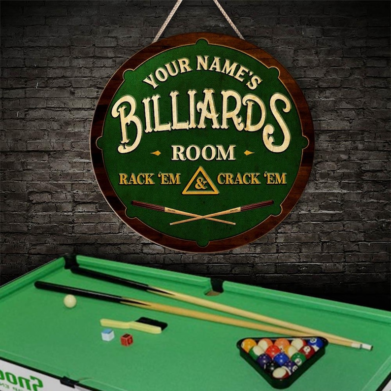May include: A round wooden sign with the text "YOUR NAME'S BILLIARDS ROOM" in a decorative font. The sign has a green center with a pool table design and the words "RACK 'EM & CRACK 'EM". A pool table with balls and cues is in the foreground.