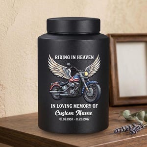 May include: A matte black cylindrical cremation urn featuring a graphic of a motorcycle with large white wings and an American flag design. The text reads RIDING IN HEAVEN, IN LOVING MEMORY OF, Custom Name, and the dates 01.08.1957 - 11.26.2017.