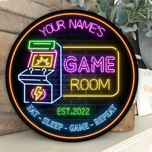 May include: Round neon sign with a black background and a brick pattern. The sign says "YOUR NAME'S GAME ROOM" with an arcade machine graphic. The sign also says "EAT - SLEEP - GAME - REPEAT" and "EST. 2022".