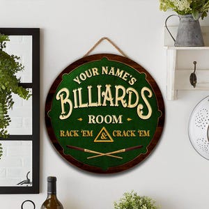 May include: A round wooden sign with a green center and a dark brown border. The sign reads "YOUR NAME'S BILLIARDS ROOM" in gold lettering. Below, it says "RACK 'EM & CRACK 'EM". Two pool cues are crossed at the bottom.