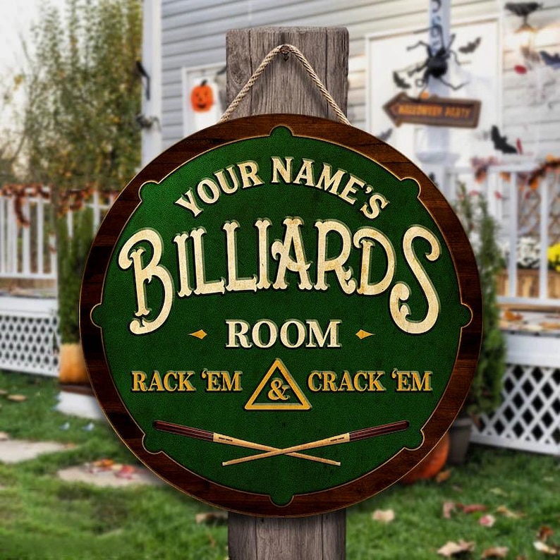 May include: A round wooden sign with a green center and a dark brown border. The sign reads "YOUR NAME'S BILLIARDS ROOM" in gold lettering, with "RACK 'EM & CRACK 'EM" below. Two crossed pool cues are at the bottom.