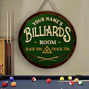 May include: A round wooden billiards room sign with the text "Your Name's Billiards Room" in white lettering. The sign has a green center and a brown border, with the phrases "Rack 'Em & Crack 'Em" below. A white rope is used for hanging.
