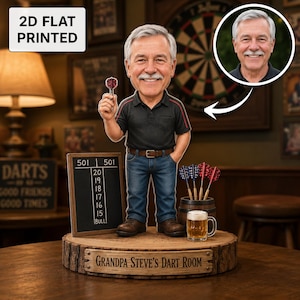 May include: A 2D flat printed bobblehead figure of a man with gray hair and a mustache, wearing a black polo shirt and blue jeans. The figure holds a dart, stands next to a dart scoreboard, a barrel of darts, and a beer mug on a wooden base.