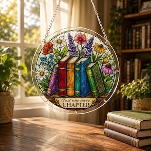 May include: A round, stained-glass window hanging with a floral and book design. The window features colorful flowers and a row of books with the text "Just one more CHAPTER". The window is suspended by a silver chain.