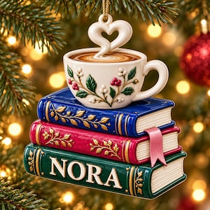 May include: A hanging ornament featuring a stack of three books in blue, red, and green with gold leaf details. The bottom book is labeled NORA in white letters. A white ceramic coffee cup with a heart-shaped steam design sits on top.