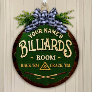 May include: Round wooden billiards room sign with a green felt center and a dark brown border. The sign features the text "Your Name's Billiards Room" and "Rack 'em & Crack 'em". It is decorated with a black and white checkered bow and greenery.