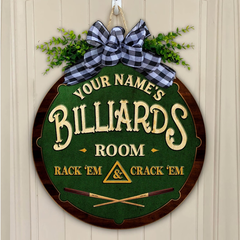 May include: Round wooden billiards room sign with a green felt center and a dark brown border. The sign features the text "Your Name's Billiards Room" and "Rack 'em & Crack 'em". It is decorated with a black and white checkered bow and greenery.