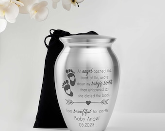 May include: A silver urn with a lid, featuring engraved text and baby footprints. The text reads "An angel opened the book of life, wrote down my baby's birth...Too beautiful for earth, Baby Angel 05.2023". A black velvet bag is next to the urn.