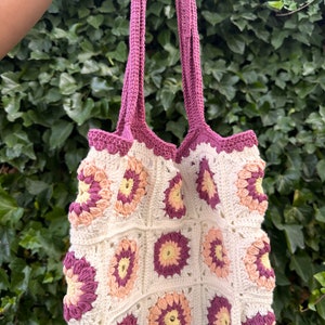 Puede incluir: Una bolsa de mano de crochet con fondo blanco y diseños florales de color rosa, melocotón y amarillo. La bolsa tiene dos asas largas de color rosa.