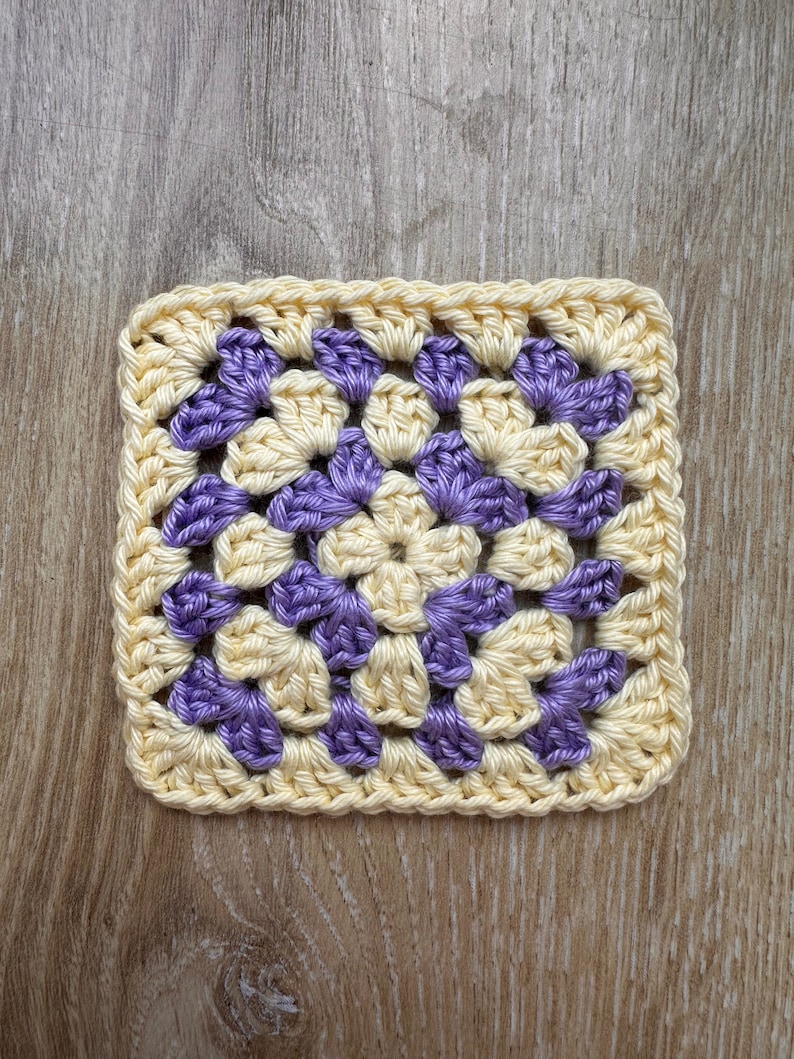 May include: A crocheted square with a yellow and purple granny stitch pattern. The square is made with a light yellow yarn and a purple yarn.