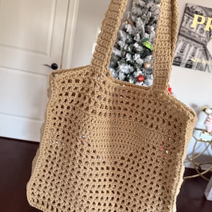 May include: A tan crochet tote bag with a rectangular body and two sturdy handles. The bag features an open, airy weave design. The bag is held up in the air.