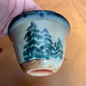 Puede incluir: Una taza de cerámica hecha a mano con una escena de bosque. La taza presenta un exterior beige claro, un borde azul y una base marrón. La escena del bosque está pintada en tonos verdes y azules.