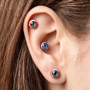 May include: Three stud earrings shaped like realistic eyeballs with bloodshot white sclera and iris details. Two earrings feature green irises with vertical pupils, while the center earring displays a blue iris with a vertical pupil.