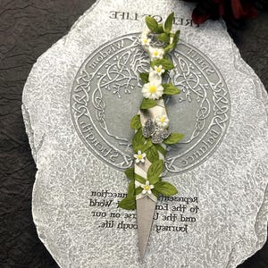 Op de afbeelding: Een decoratieve dolk met een zilveren lemmet, versierd met witte madeliefjes, groene bladeren en een zilveren vlinder. Het handvat is omwikkeld met wit lint. De dolk ligt op een grijze steen met gegraveerde tekst en een boomontwerp.