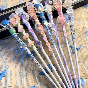 Puede incluir: Una colección de ocho varitas decorativas con diseños ornamentados. Cada varita presenta una combinación única de colores, incluyendo azul, rosa, morado y blanco, con adornos de cristal y flores. Las varitas se exhiben sobre un fondo de periódico vintage.