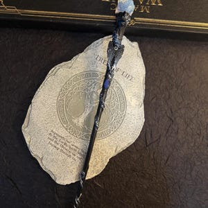 Puede incluir: Una varita m&aacute;gica artesanal con una punta de cristal azul, intrincadas tallas de madera oscura y una base en espiral plateada. La varita descansa sobre una piedra con las palabras "Tree of Life" y otro texto. La marca "HUEHANDIWORK" es visible.