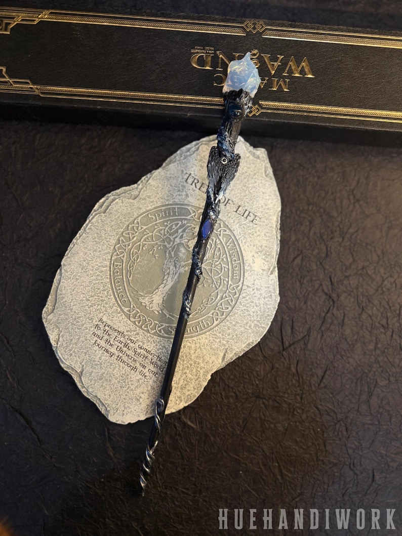 Puede incluir: Una varita m&aacute;gica artesanal con una punta de cristal azul, intrincadas tallas de madera oscura y una base en espiral plateada. La varita descansa sobre una piedra con las palabras "Tree of Life" y otro texto. La marca "HUEHANDIWORK" es visible.