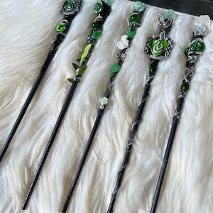 Könnte beinhalten: Fünf schwarze Zauberstäbe mit dekorativen Griffen. Die Zauberstäbe weisen grüne und silberne Akzente auf, darunter Edelsteine, Blätter und florale Designs. Jeder Zauberstab hat ein einzigartiges Griffdesign, das auf einer weißen, flauschigen Oberfläche liegt.