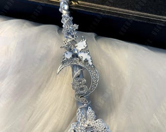 May include: A silver-colored wand with intricate detailing. The wand features a clear crystal orb at the base, a crescent moon design, and a snake wrapped around the shaft. White floral and crystal accents adorn the wand.