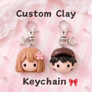 May include: Two custom clay keychains featuring stylized character heads with black eyes. One has light brown hair with a pink flower, the other has dark brown hair and a cap. Both have silver lobster clasps with star and moon charms. Text reads Custom Clay Keychain.