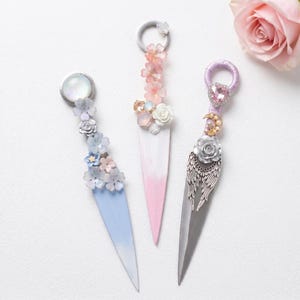Op de afbeelding: Drie decoratieve briefopeners met sierlijke handvatten. Eén heeft een blauw-wit gekleurd lemmet met bloemaccenten. Een andere heeft een roze-wit gekleurd lemmet met roze bloemen. De derde heeft een zilveren lemmet met een rozen- en vleugelontwerp.