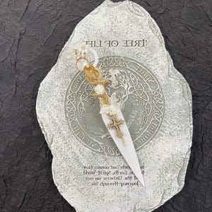 May include: A white stone with the words "Tree of Life" inscribed on it. A white and gold decorative dagger with a cross is placed on the stone. The stone has a Celtic knot design around the edge.