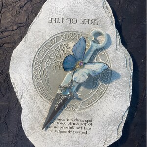 May include: A white stone with a silver circle in the center. The circle has a tree of life design with a blue butterfly and a silver key. The text "TREE OF LIFE" is written above the circle. The text "Represents our journey through life and the universe. Spirit on our to the earth.  " is written below the circle.
