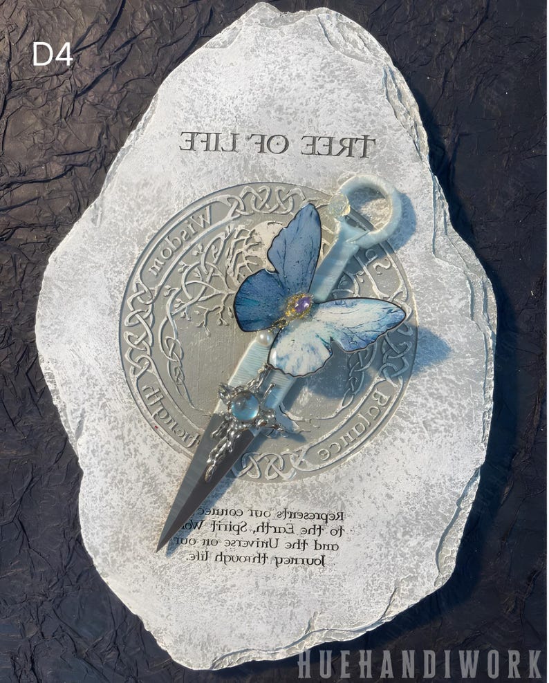 May include: A white stone with a silver circle in the center. The circle has a tree of life design with a blue butterfly and a silver key. The text "TREE OF LIFE" is written above the circle. The text "Represents our journey through life and the universe. Spirit on our to the earth.  " is written below the circle.