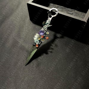 Puede incluir: Una daga decorativa con mango plateado y hoja verde oscuro. El mango está adornado con flores coloridas, una gema púrpura y un anillo plateado. El fondo es negro.