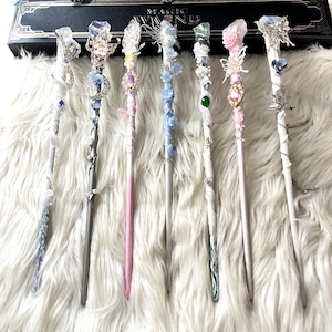 May include: Seven decorative magic wands with crystal and floral details. Each wand has a unique design with colours like pink, blue, and silver. The wands are displayed on a fluffy white surface, with a box labelled "MAGIC WAND" in the background.