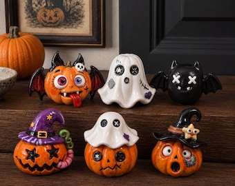 May include: A collection of six ceramic Halloween figurines including three orange pumpkins with expressive faces, one white ghost, one black bat with white eyes, and one pumpkin wearing a purple witch hat, all featuring detailed painted accents.