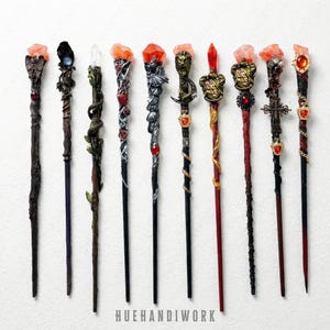May include: A collection of nine intricately designed wands. Each wand features unique embellishments, including crystals, sculpted figures, and decorative accents. The wands vary in color, with details in silver, gold, red, and black. The text "HUEHANDIWORK" is at the bottom.