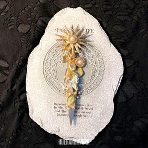 以下が含まれることがあります： 生命の樹のデザインが施された装飾用の石。金色のサンバースト、真珠、葉のアクセントが特徴です。石にはエッチングされたテキストと中央のデザインがあります。全体的なカラースキームは金、白、グレーです。