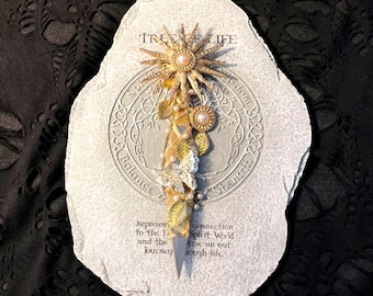May include: A decorative stone with a Tree of Life design, featuring a gold sunburst, pearls, and leaf accents. The stone has etched text and a central design. The overall color scheme is gold, white, and gray.