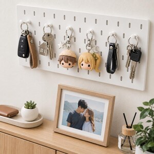 May include: A white rectangular pegboard wall organizer with six white pegs holding various keychains, including a car key fob, a house-shaped charm, two stylized character heads with hair, and sets of metal keys with leather straps.