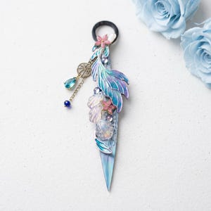 May include: A mermaid-themed hair accessory with a black elastic band. The design features a blue and purple iridescent tail, pink flowers, and a dangling charm with blue and teal gems. The accessory is embellished with seashells and crystals.