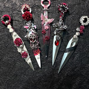 May include: Five decorative kunai knives with ornate handles and blades. Each knife features unique embellishments, including roses, butterflies, and crosses. The handles are adorned with pearls, gems, and metal chains. The blades are silver and the overall color scheme includes red, silver, and black.