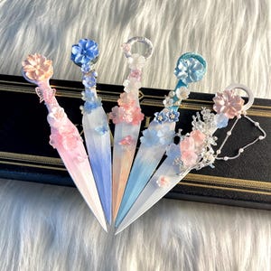 以下が含まれることがあります： パステルカラーの刃と花の装飾が施された、装飾的な花のレターオープナー5つ。 オープナーは、白い毛皮の背景を持つ黒い表面に配置されています。 刃はピンク、青、白の色合いで塗装されています。