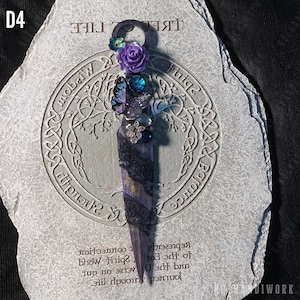 May include: A purple and blue decorative dagger with a flower and butterfly design. The dagger is on a gray stone with the words "Tree of Life" and "Spirit Within" inscribed on it.