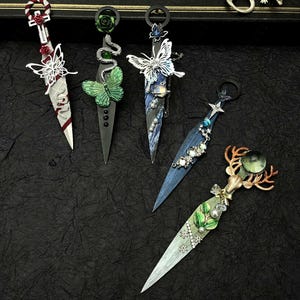 May include: Five decorative fantasy daggers with ornate handles and blades. The daggers feature butterfly, snake, and deer designs, with accents of red, green, and silver. The text "MAGIC WAND" is visible on a black box in the background.