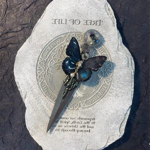May include: A gray stone with the words "Tree of Life" inscribed on it. A black and blue butterfly with a pearl accent is attached to a silver knife. The knife is placed on a circular design with the words "Balance", "Wisdom", and "Truth" inscribed around it.