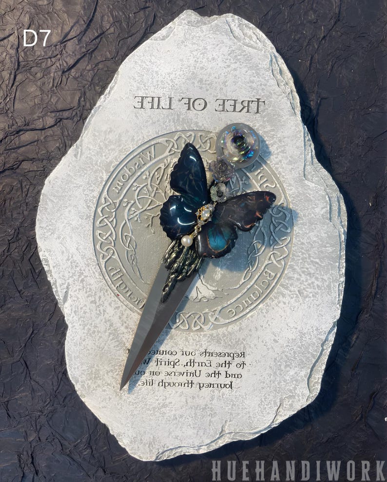 May include: A gray stone with the words "Tree of Life" inscribed on it. A black and blue butterfly with a pearl accent is attached to a silver knife. The knife is placed on a circular design with the words "Balance", "Wisdom", and "Truth" inscribed around it.