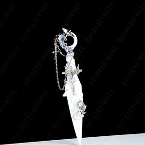 May include: A decorative dagger with a white blade and ornate silver handle. The handle features a crescent moon, chains, and various gemstones. The blade is adorned with a cross and other embellishments. The background is black and white.