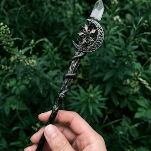 May include: A black wand with a crystal tip, a crescent moon, and a star design. The wand has a snake wrapped around the shaft. The wand is held against a backdrop of green foliage.